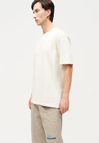 Jeune homme portant un T-shirt uni blanc cassé et un pantalon cargo beige, debout devant un fond neutre.