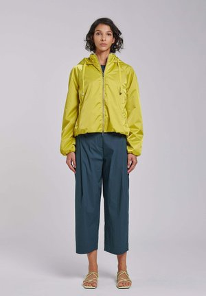 Kobieta stojąca w limonkowozielonej bluzie z kapturem na suwak, ciemnych szerokich spodniach o długości do połowy łydki i lekkich zielonych sandałach na paski na jednolitym tle.