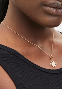 Rosen-guld halskæde med en rektangulær pendants, som har et præget design, hængende på en fin kæde mod en mørk, ribbet top.