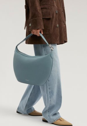 Personne en manteau marron et jean bleu clair tient un sac à main en cuir bleu incurvé, portant des chaussures pointues beiges.