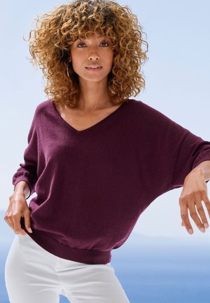 Woman with curly hair wearing a purple V-neck sweater and white pants, posing with one hand on hip and ocean background.