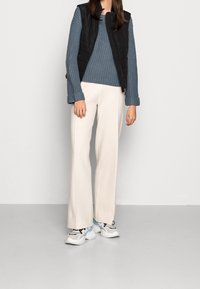 Kvinna som bär en blå stickad tröja, svart vadderad väst, krämfärgade vida byxor och vita klumpiga sneakers står mot en enfärgad vägg.