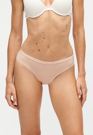 Kvinde iført en hvid bh og lyse beige bikiniunderbukser, stående med armene afslappet langs siden mod en ensfarvet baggrund.
