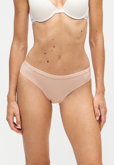 Femme portant un soutien-gorge blanc et une culotte bikini beige clair, debout, les bras détendus le long du corps, sur un fond uni.