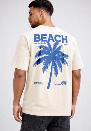 Mann von hinten gesehen, trägt ein beiges T-Shirt mit einer großen blauen Palme und dem Schriftzug „BEACH Social Club 1976“ auf dem Rücken.