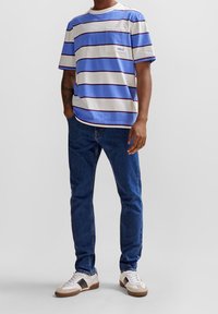 Pruhované tričko s krátkým rukávem v světle modré, bílé a tmavě modré barvě, s přední kapsou a logem. Nošeno s modrými džíny a teniskami.