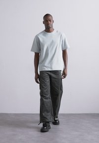 Lyseblå bomulds-t-shirt med korte ærmer, parret med mørkegrønne cargo-bukser. Modellen har sorte klodsede støvler på og står foran en neutral baggrund.