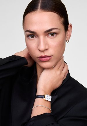 Montre carrée avec un cadran blanc, un bracelet en cuir noir et des éléments en or. Portée avec un bracelet délicat en argent. Le modèle porte un blazer noir.