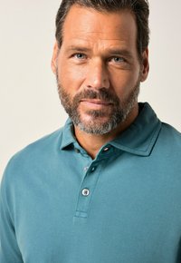 Turkusowa koszulka polo z teksturowanej tkaniny, z kołnierzykiem i zapięciem na dwa guziki. Zbliżenie, podkreślające detale szwów i guzików.