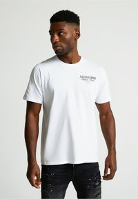 Biely bavlnený tričko s krátkymi rukávmi, okrúhlym výstrihom a malým potlačeným logom na ľavej strane hrudníka. Nosené s čiernymi roztrhnutými džínami.