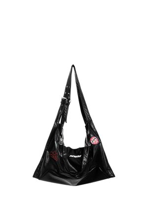 Joven con cabello trenzado que lleva una chaqueta de cuero gráfica en blanco y negro, pantalones negros con logo verde y zapatos negros texturizados.