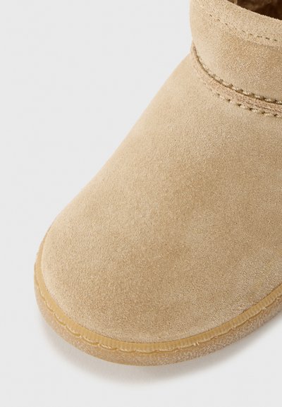 Beige semsket ankelstøvler med avrundet tå, med en sydd bånd rundt kragen og gummisåle. Myk tekstur og minimalistisk design.