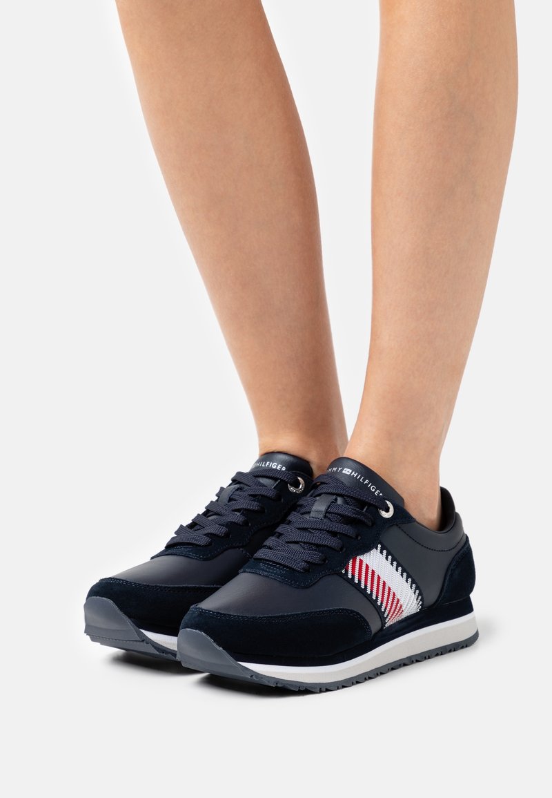 Ben som bär svarta sneakers med vita, röda och marinblå stickade ränder och svarta snören mot vit bakgrund.