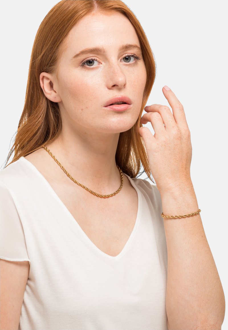 Collier et bracelet en chaîne tordue dorée sur une personne. Le collier s'ajuste étroitement autour du cou, le bracelet est porté au poignet.