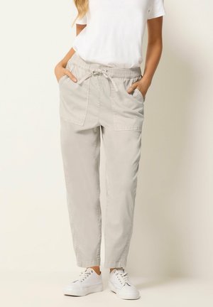 Femme portant un pantalon beige clair avec cordon de serrage et poches avant, un t-shirt blanc et des baskets blanches, debout les mains dans les poches.