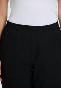 Zwarte joggingbroek met een elastische tailleband, een gladde textuur en zijzakken, naast een witte tanktop gepresenteerd.