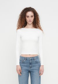 Λευκό crop top μακρυμάνικο με λαιμόκοψη σκάφος, συνδυασμένο με ανοιχτόχρωμα γαλάζια ψηλόμεσα τζιν, που παρουσιάζει εφαρμοστή γραμμή και ομαλή υφή.
