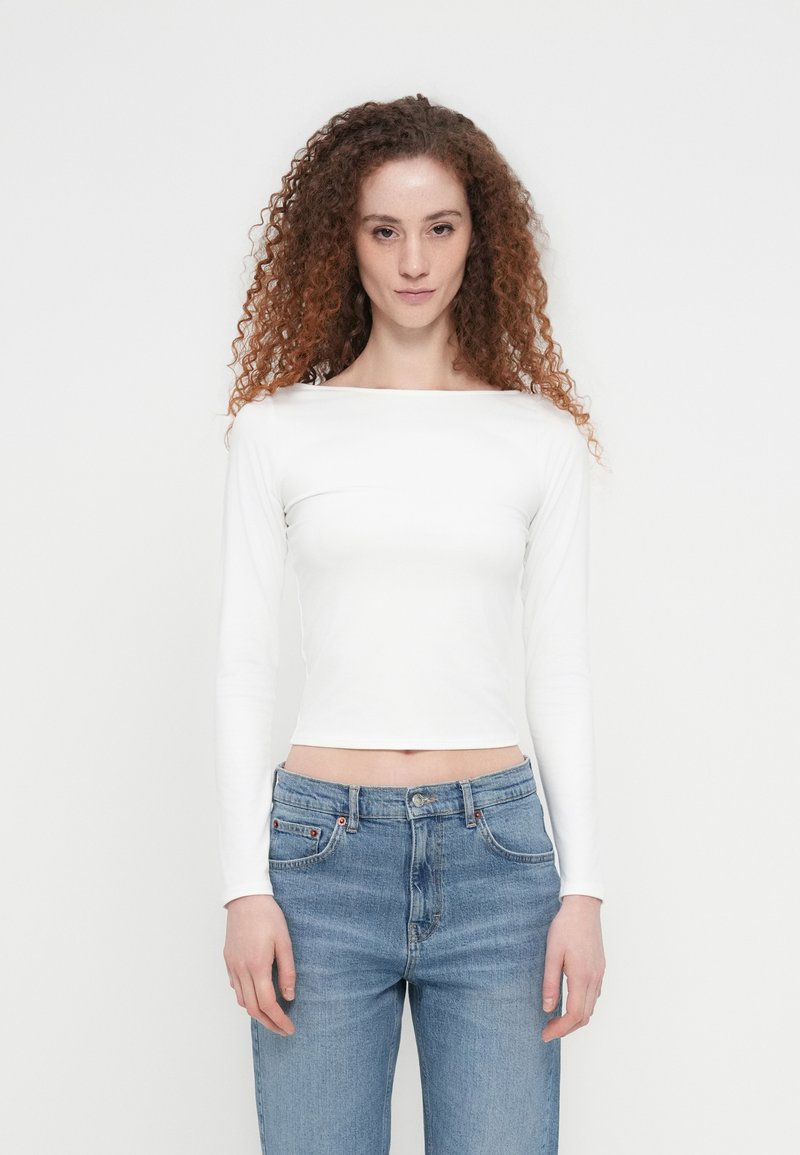 Λευκό crop top μακρυμάνικο με λαιμόκοψη σκάφος, συνδυασμένο με ανοιχτόχρωμα γαλάζια ψηλόμεσα τζιν, που παρουσιάζει εφαρμοστή γραμμή και ομαλή υφή.