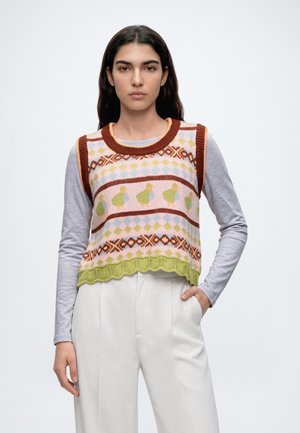 Le modèle porte un gilet en maille coloré à motifs sur un haut gris, un pantalon blanc à jambes larges, des chaussures plates bordeaux et un cabas marron foncé.