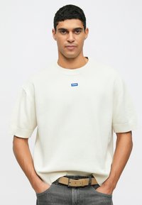 Mann med kort, krøllete svart hår iført en hvit kortermet genser med en liten blå "HUGO"-logo, grå jeans og et beige belte, med hendene i lommene.