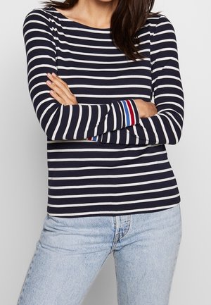 Maglietta a righe blu navy e bianco con maniche lunghe e scollo arrotondato, decorata con accenti in rosso, bianco e blu ai polsini. Abbinata a jeans azzurri.