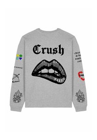 Pelēks krekls ar lielu melnu lūpu grafiku un tekstu "Crush" augšpusē. Dažādi krāsaini plāksteri uz piedurknēm ar tekstu un ikonām.
