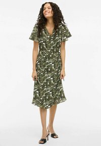 Zelené květinové šaty s potiskem listů, krátkými volánkovými rukávy a střední délkou. Nosič má na sobě černé sandály na otevřenou špičku.