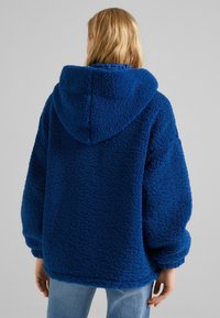 Luddig blå hoodie i tjockt, texturerat fleece-material, med huva och oversized passform. Enkel design, utan synliga mönster eller detaljer.
