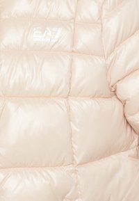 Jasny beżowy pikowany płaszcz z prostokątnym przeszyciem i srebrnym logo EA7 Emporio Armani na piersi.