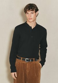 Schwarzes Langarm-Henley-Shirt mit lockerer Passform, ausgestattet mit einer Knopfleiste und kombiniert mit braunen Cordhosen, die mit vertikalen Streifen strukturiert sind.