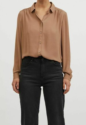 Kobieta ubrana w koszulę na guziki z długim rękawem w kolorze wielbłądzim, włożoną do wysokowarstwowych czarnych dżinsów, stojąca na jednolitym tle.