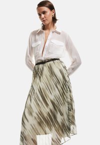 Camisa blanca de manga larga con bolsillos en el pecho, combinada con una falda plisada y transparente que presenta patrones abstractos en verde y beige, y un cinturón negro.