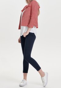 Rode gebloemde cropped jack met gerimpelde randen over een witte top, gecombineerd met navy skinny jeans en witte sneakers.