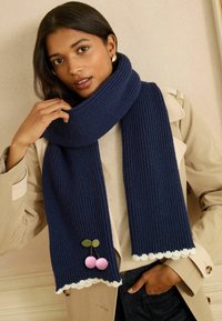 Mørkeblå ribstrikket tørklæde med bølget cremefarvet kant og pink kirsebær pom-pom dekorationer; båret omkring halsen.