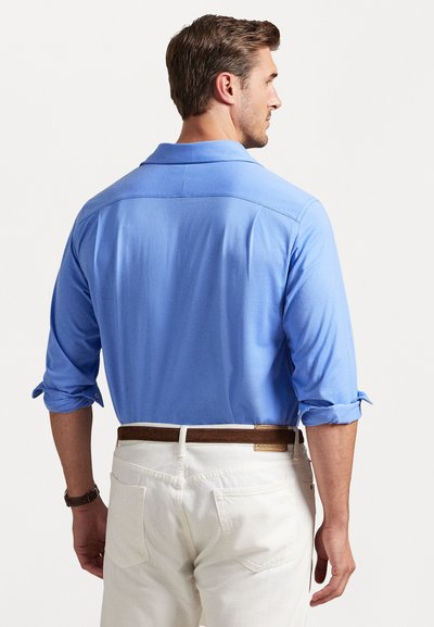 Ανοιχτό μπλε μακρυμάνικο πουκάμισο με γιακά και αναδιπλωμένα μανίκια, με απαλό �υφή. Φοριέται με λευκά παντελόνια και καφέ ζώνη.