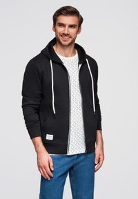 Felpa nera con zip e coulisse bianche, indossata sopra una camicia bianca a fantasia. Presenta tasche frontali e un'etichetta in tessuto con logo. I jeans blu completano l'outfit.