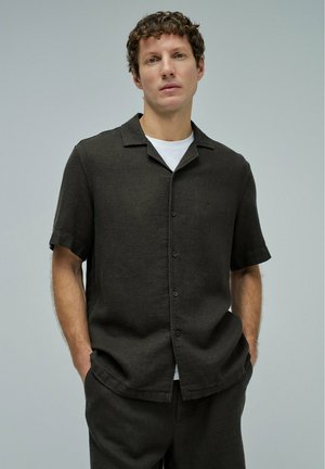 Hombre con cabello rizado que lleva una camisa oscura de manga corta abotonada sobre una camiseta blanca, con las manos en los bolsillos, frente a un fondo claro y liso.