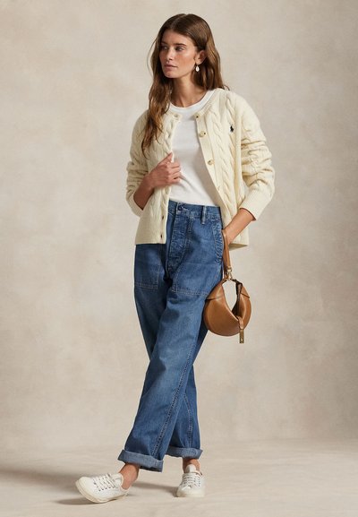 Femeie purtând un cardigan crem cu model împletit peste o cămașă albă, blugi albaștri cu talie înaltă, adidași albi, ținând o geantă de mână maro din piele pe lângă ea.