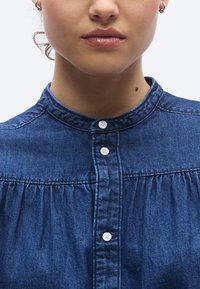 Das Jeanshemd verfügt über einen Kragen, eine vordere Knopfleiste mit weißen Knöpfen und Raffdetails unterhalb der Brust in einem tiefen Blauton.