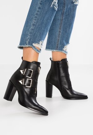 Schwarze hohe Leder-Stiefeletten mit doppelten silbernen Schnallen, getragen zu kurz geschnittenen, zerrissenen blauen Jeans, vor einem weißen Hintergrund.