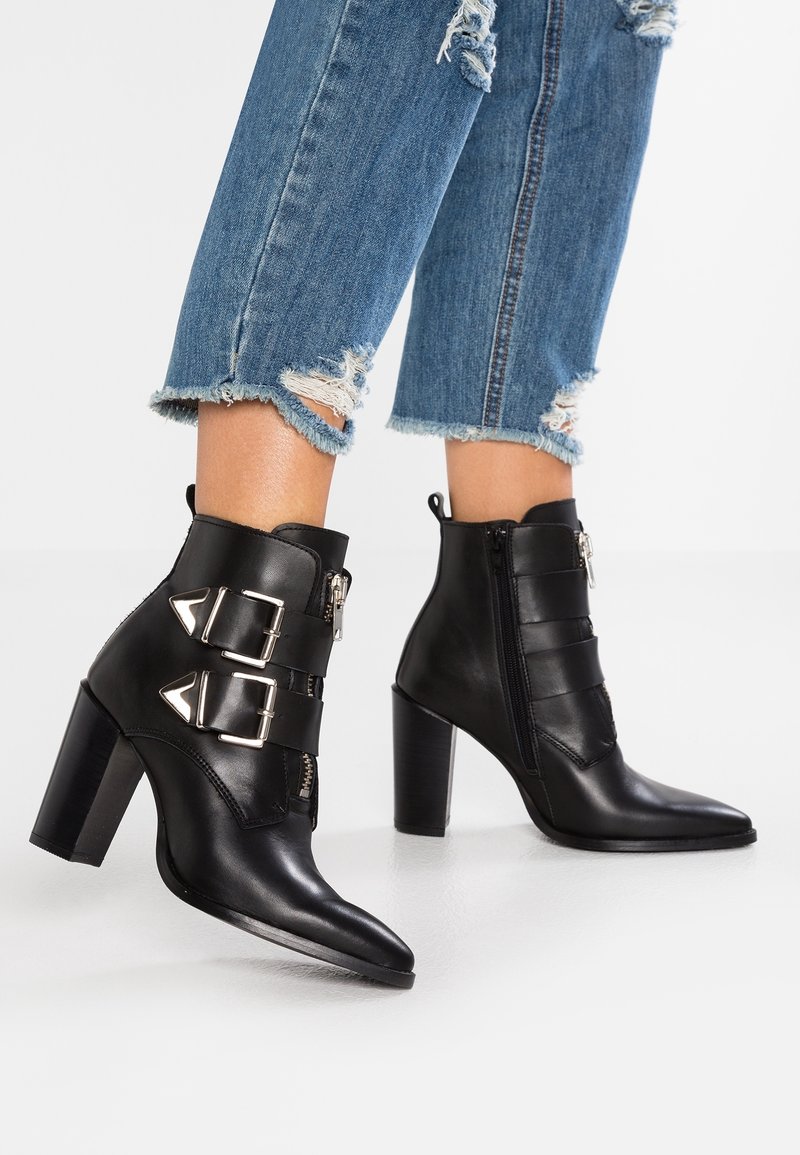 Bottes chevilles en cuir noir à talons hauts avec deux boucles argentées, portées avec un jean bleu déchiré et raccourci sur un fond blanc uni.