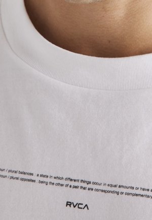 Gros plan sur le col d'un t-shirt RVCA blanc avec un texte descriptif sur les "équilibres" imprimé au-dessus du logo de la marque.