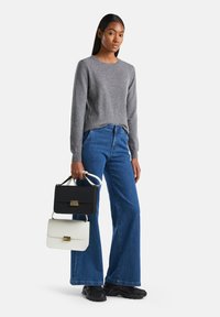 Kobieta w szarym swetrze i niebieskich dżinsach trzyma czarne i białe torebki, stojąc na tle jednolitego białego tła.