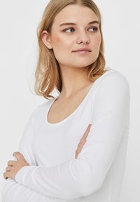Witte lange mouwen top met een ronde hals, gemaakt van zachte stof. Het model heeft de armen gekruist, waardoor de gladde textuur en pasvorm zichtbaar zijn.