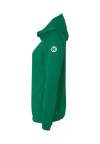 Grøn softshell-jakke med hætte, der har en lynlåslukning foran, sidelommer og et cirkulært logo på venstre arm. Glat tekstur.