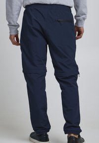 Pantalon décontracté bleu marine avec taille élastique, poche arrière zippée et jambes fuselées. Confectionné dans un tissu léger à la texture lisse.
