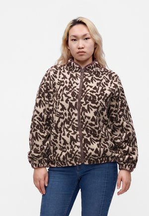 Ung kvinde med blondt hår iført en brun og beige mønstret fleecejakke med lynlås og blå jeans mod en ensfarvet baggrund.