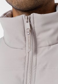 Jachetă gri deschis, cu guler înalt, material neted, fermoar frontal cu branding și detalii de cusătură contrastante.