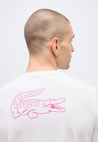 Άνδρας με κοντά μαλλιά και σκουλαρίκι που φοράει λευκό πουκάμισο με μεγάλο ροζ κροκόδειλο στο πίσω μέρος, γυρισμένος πλάτη στην κάμερα.