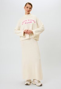 Crème-kleurige gebreide trui met "GOOD KARMA CLUB" in roze letters, gecombineerd met een bijpassende midi-rok en gedragen met beige sneakers.
